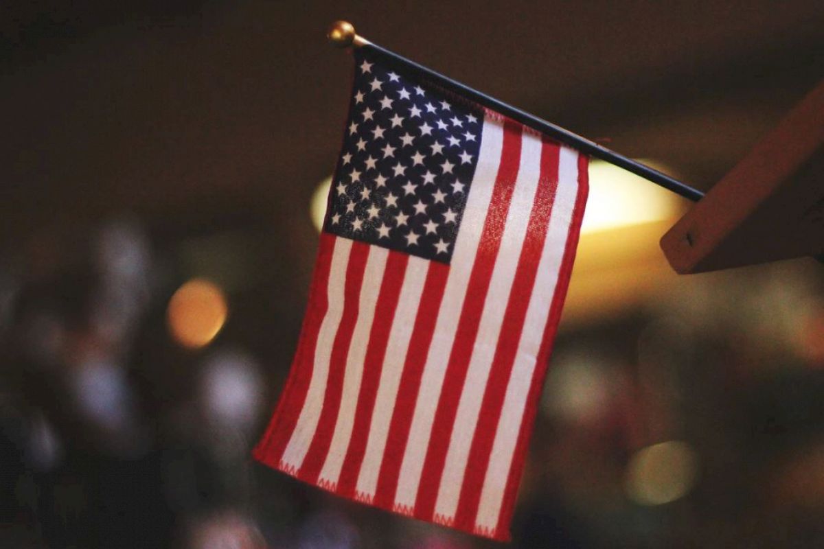 The image shows a small United States flag with 13 stripes and 50 stars, mounted on a pole with a blurred background.