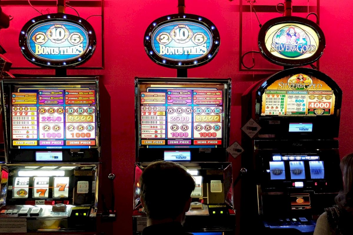 The image shows three people playing on slot machines in a casino with colorful screens displaying various game options and rewards.