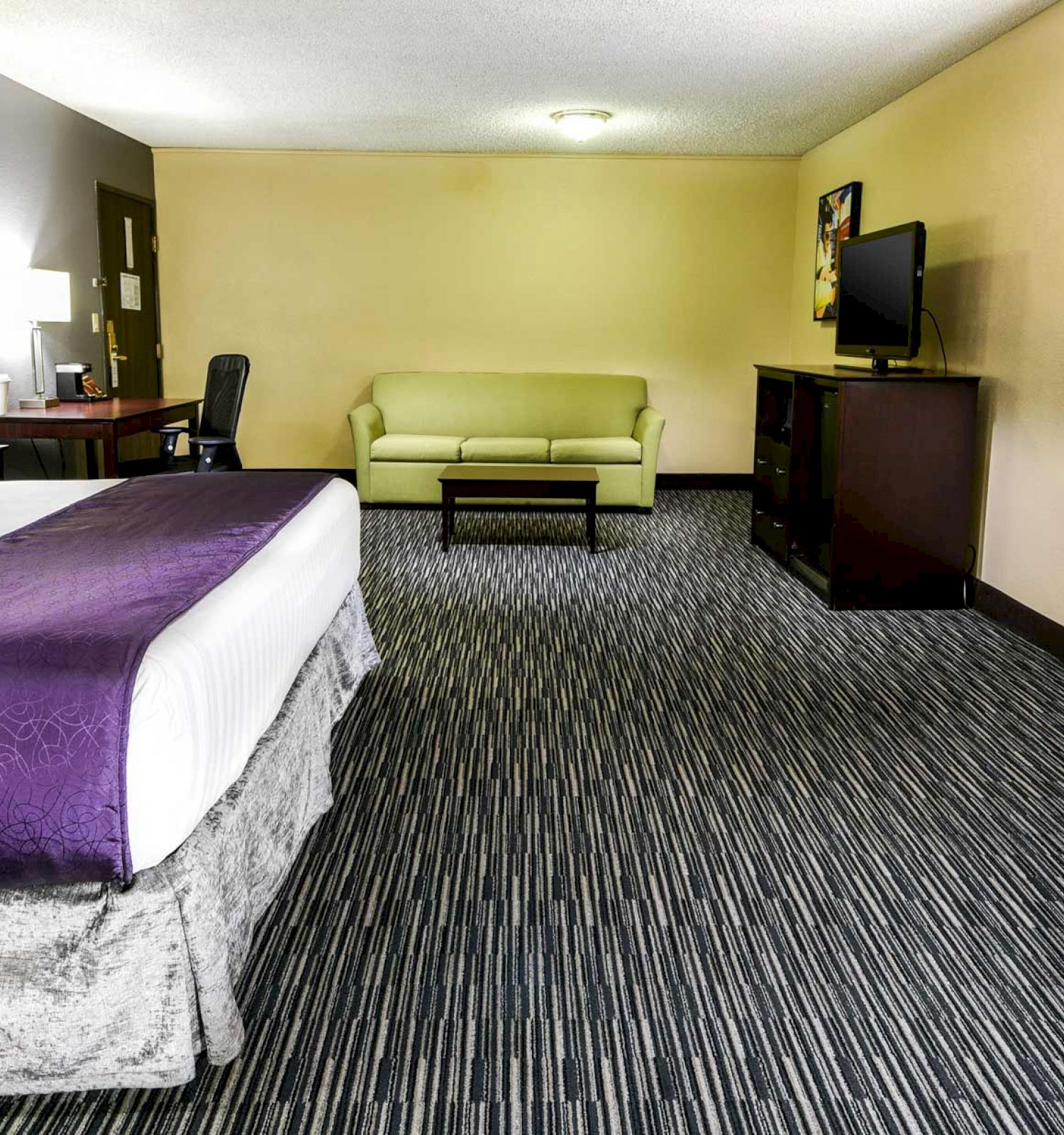 A hotel room with a king bed, purple throw, desk, TV, green sofas, air conditioner, and modern decor.