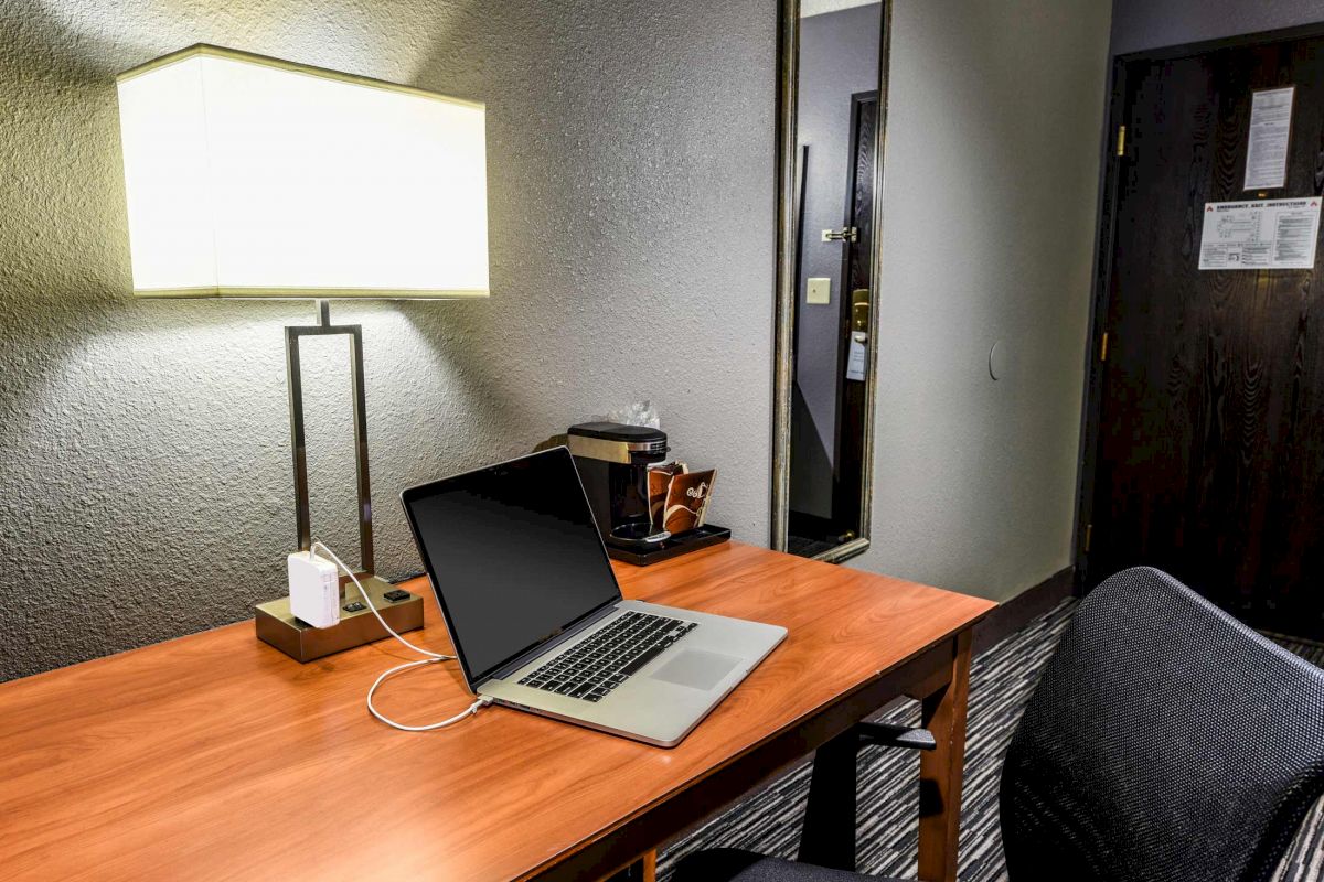 A hotel room desk with a lamp, laptop, coffee maker, and mirror on the wall, with the door visible in the background, creating a tidy workspace.