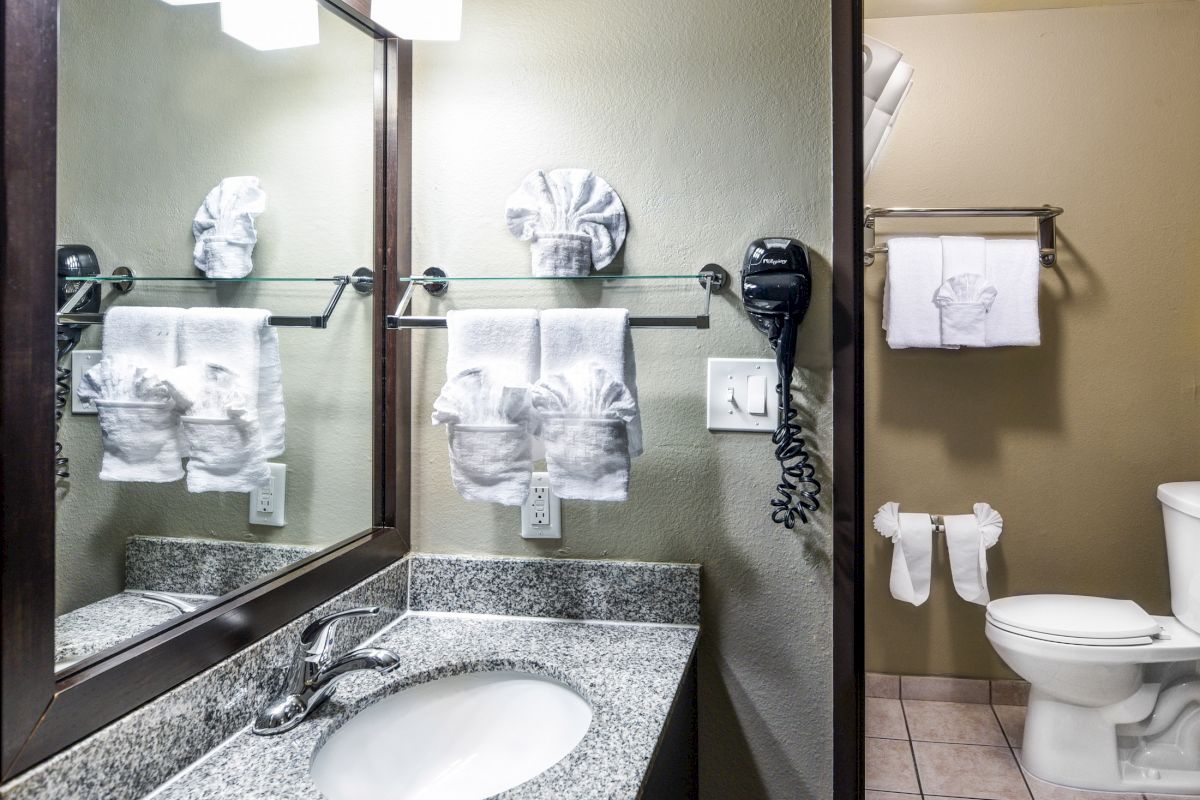 A clean bathroom featuring a granite countertop with a sink, mirror, towels, a hairdryer, and a toilet in the background.