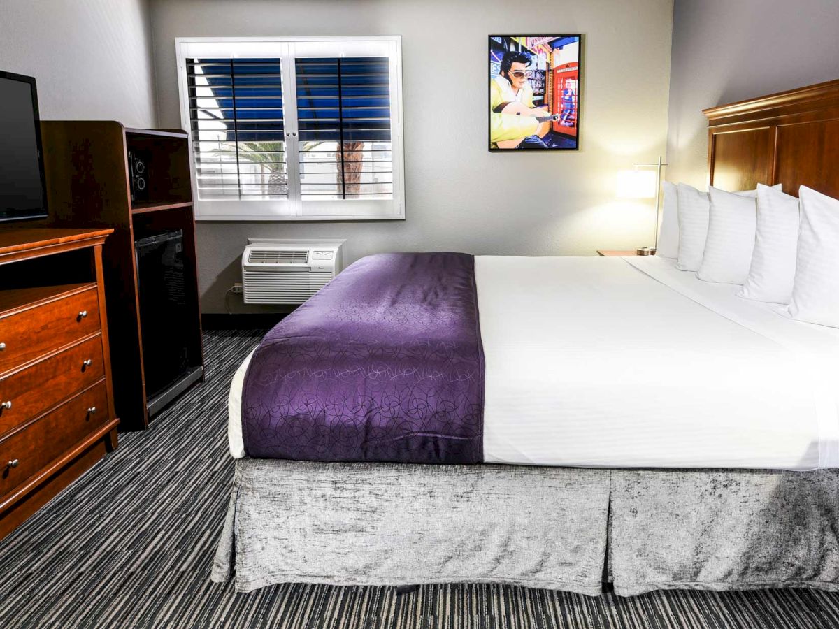 A hotel room with a large bed, a TV on a dresser, a small fridge, an air conditioner, and a window. There's a wall picture and a lamp.