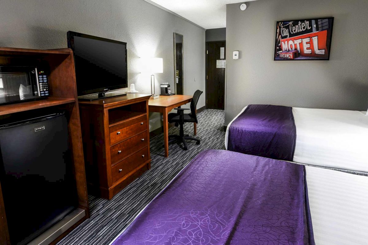 The image shows a hotel room with two beds, a TV on a dresser, a mini-fridge, a desk with a chair, and a wall-mounted sign reading 