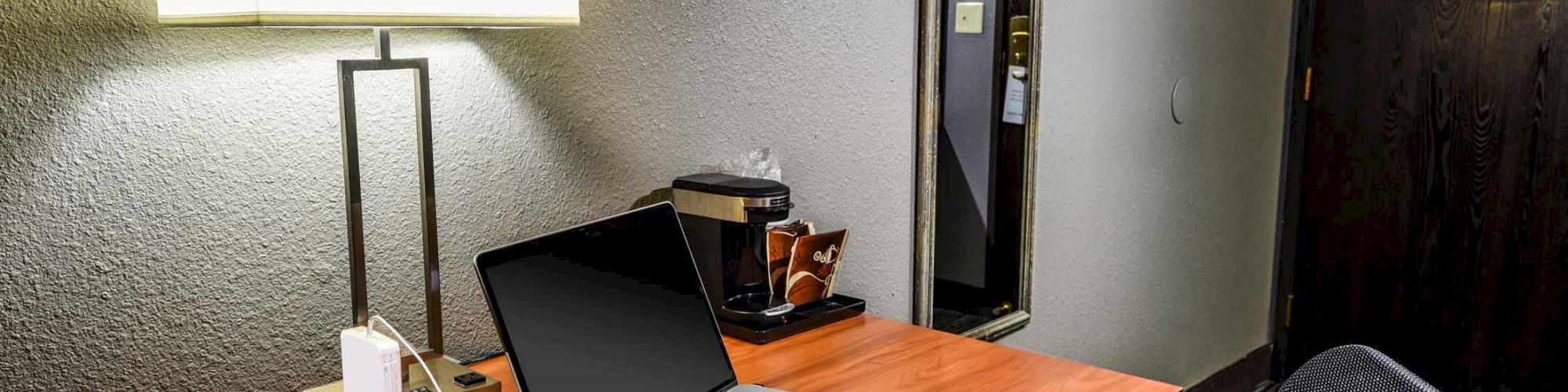 A desk with a lamp, an open laptop, a chair, and a coffee maker against a wall with a mirror and a door.