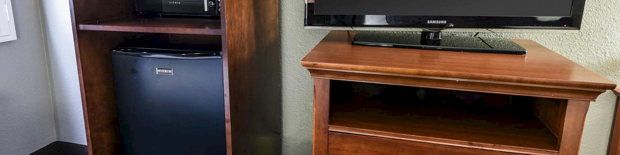 The image shows a wooden cabinet with a black microwave and mini-fridge next to a dresser with a flat-screen TV on top.