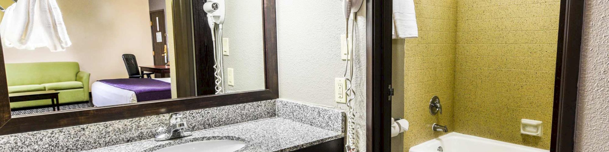 The image shows a hotel bathroom with a granite countertop, sink, mirror, hairdryer, and bathtub with a shower, next to a bedroom area.