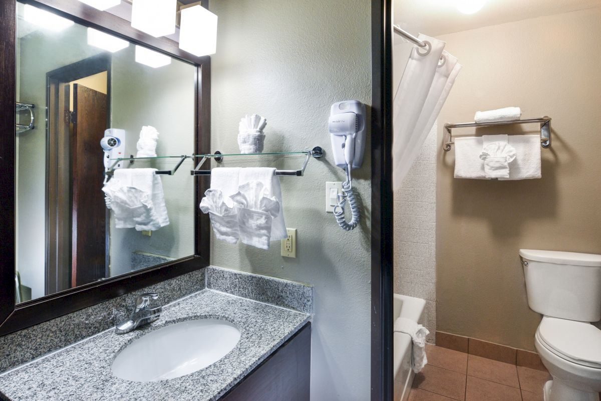 The image shows a hotel bathroom with a sink, mirror, neatly arranged towels, a hairdryer, and a toilet next to a bathtub with a shower curtain.