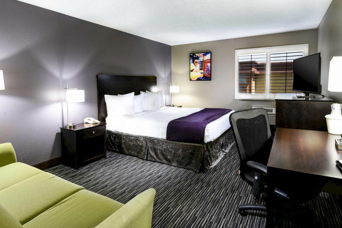 The image shows a modern hotel room with a double bed, a desk with a chair, a TV, a sofa, and decorative lamps, with a window on the wall.