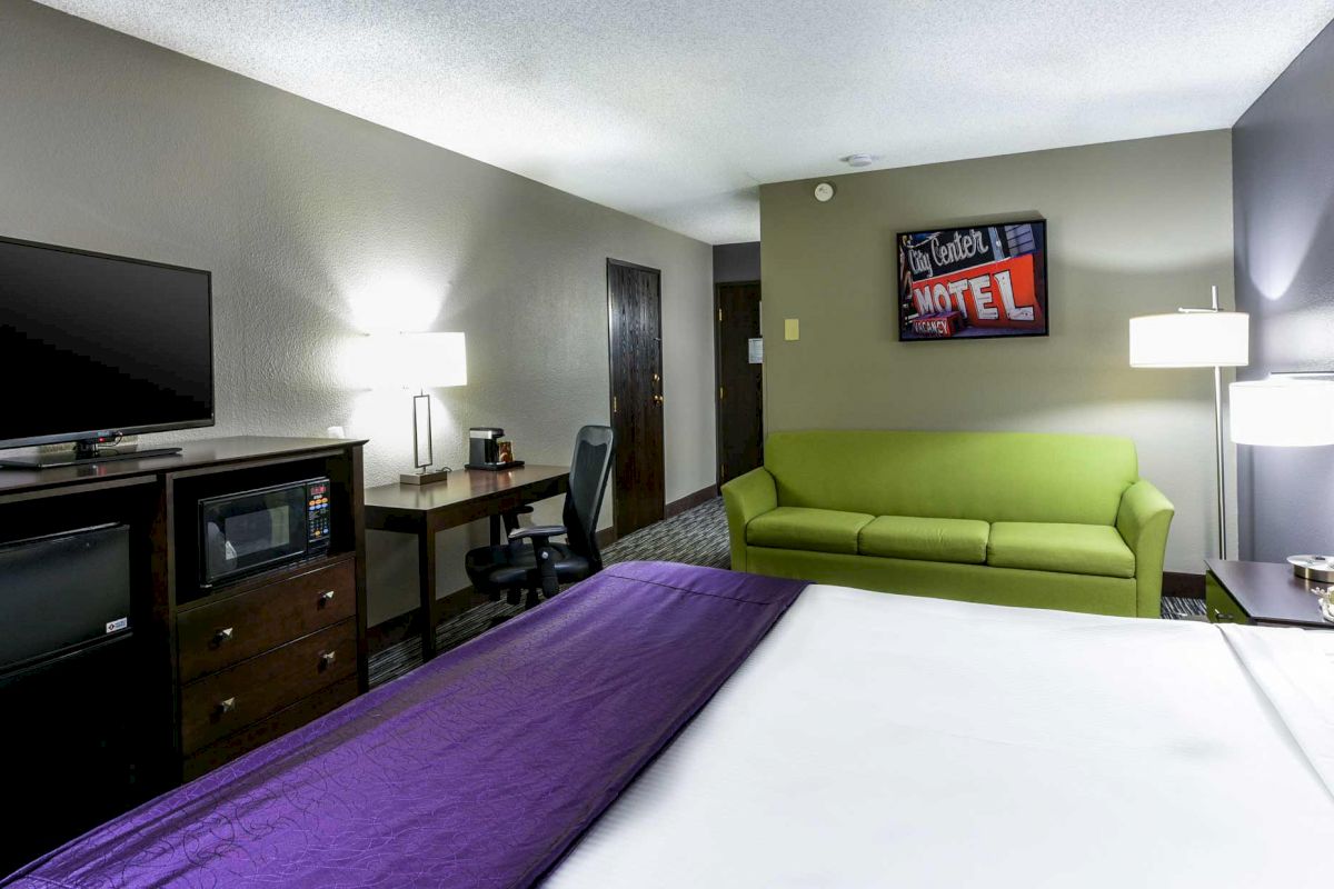 The image shows a hotel room with a bed, green sofa, desk, TV, microwave, and lamps, decorated with a Motel-themed art piece on the wall.