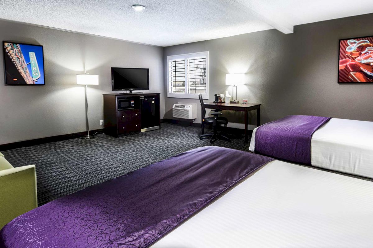 A modern hotel room with two beds covered in purple blankets, a green sofa, a desk, a TV, and colorful artwork on the walls.