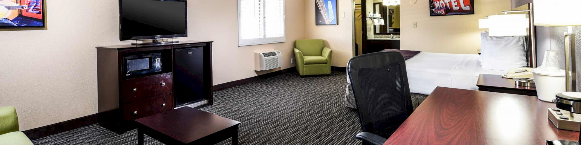 A modern hotel room with a bed, desk, chair, sofa, TV, and decor. There is a window, artwork on the walls, and a small coffee station on the desk.