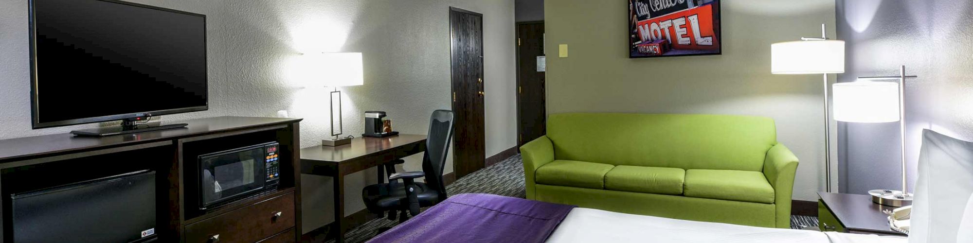 The image shows a hotel room with a bed, green sofa, TV, desk, microwave, and lamps on a nightstand and desk. A painting says 