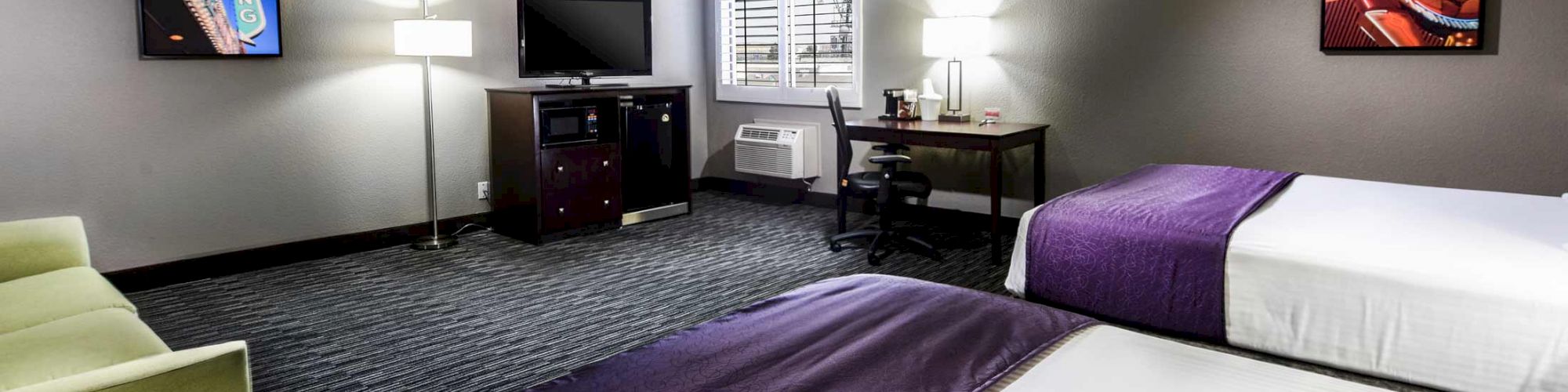 A hotel room with two purple-bedded beds, a green sofa, a desk with a chair, a TV on a stand, and two framed art pieces on the wall ends the sentence.