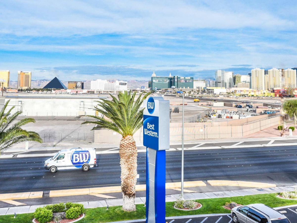 A cityscape featuring a Best Western sign, a Blu truck, palm trees, a road, and tall buildings in the background, under a blue sky.