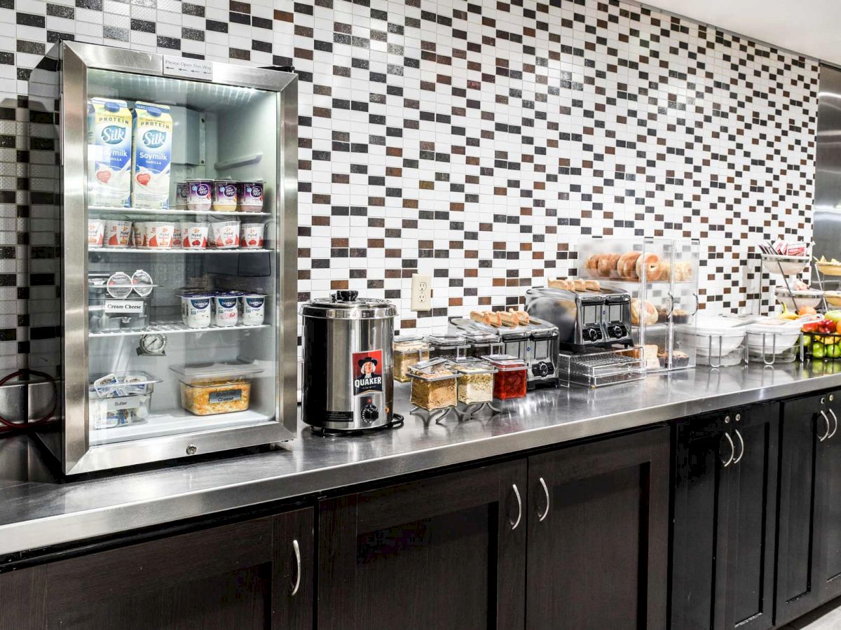 A breakfast buffet setup with juices, yogurt, coffee, bread, cereals, and fruits presented against a black and white tiled wall.