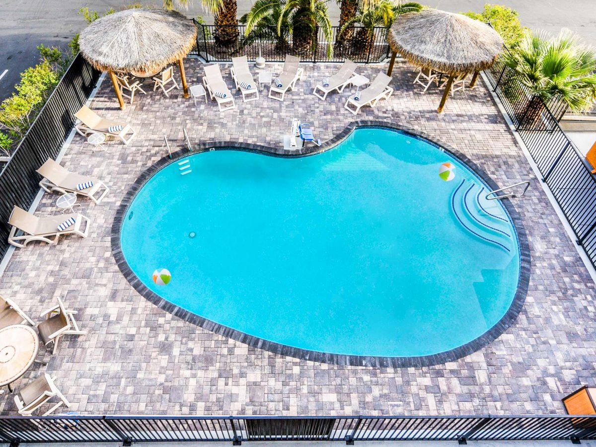 An overhead view of a heart-shaped swimming pool surrounded by lounge chairs, two thatched umbrellas, and a table with chairs, enclosed by a fence.