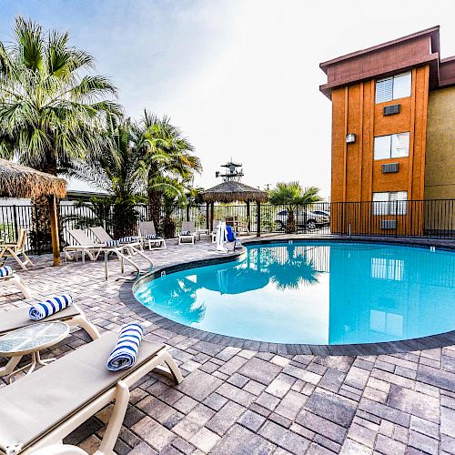 The image shows a pool area with lounge chairs, blue and white striped towels, palm trees, and a building in the background.