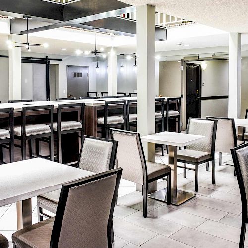 The image shows a modern, clean dining area with tables and chairs, a bar counter with stools, light fixtures, and a TV mounted on the wall.