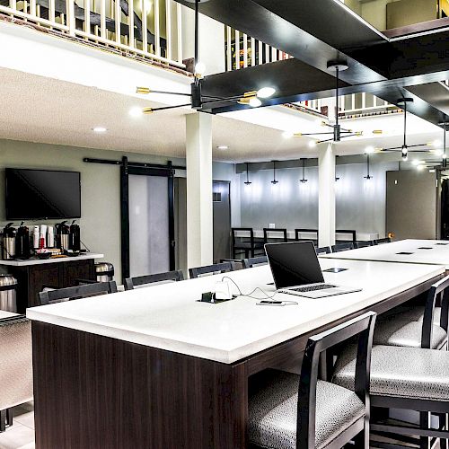 A modern communal workspace with a large table, chairs, laptop, coffee station, and TV, illuminated by stylish overhead lights.