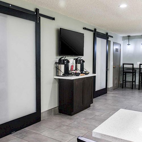 The image shows a modern room with a coffee station, a television, bar seating, and tables. Sliding doors lead to another area, all in a sleek design.