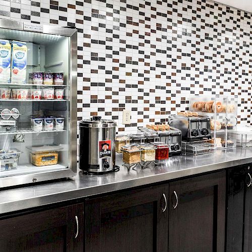 This image shows a breakfast buffet with a juice dispenser, mini-fridge with milk and yogurt, coffee machine, cereal dispensers, and various pastries.