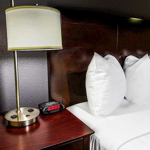 A bedside table with a lamp, digital clock showing 2:06, and a bed with white pillows and sheets next to it ending the sentence.