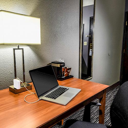 A desk with a laptop, lamp, and coffee maker in a room. The scene also includes a chair, a wall mirror, and a door in the background.