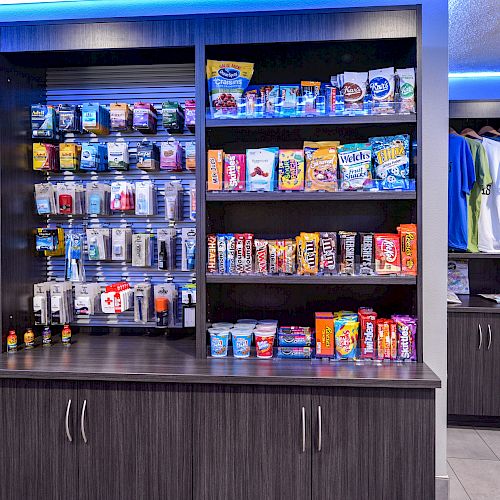 The image shows a store with snacks, beverages, toiletries, and clothing neatly arranged on wooden shelves with a blue backlight.