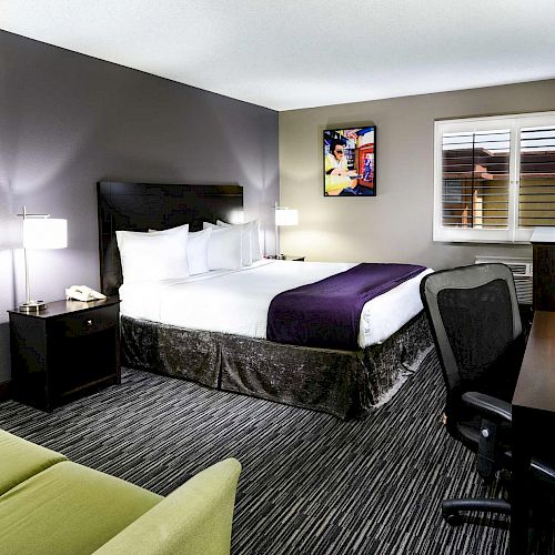 A modern hotel room features a queen-sized bed, a desk with a chair, a sofa, two bedside tables with lamps, and a wall-mounted flat-screen TV.