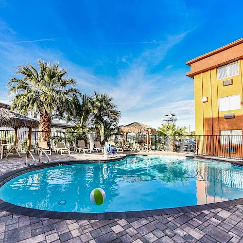 The image shows an outdoor swimming pool surrounded by lounge chairs, palm trees, and a building, with a clear blue sky overhead and some inflatables in the water.