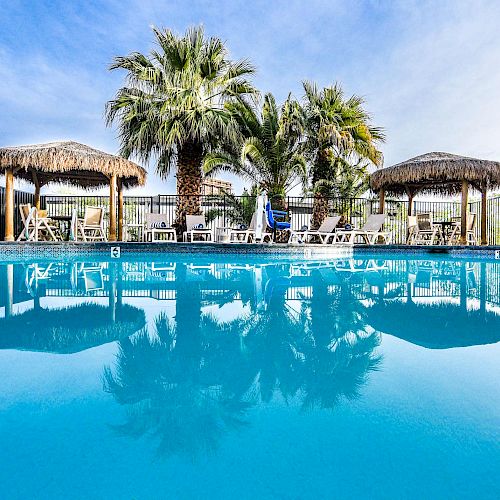 A serene swimming pool with clear blue water, palm trees, and sun loungers under thatched umbrellas, set against a bright sky.