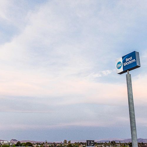 The image shows a Best Western hotel sign on a tall pole against a cloudy sky, with a distant cityscape and communication tower below it.