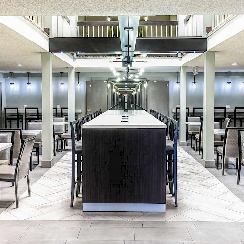 The image shows a spacious dining hall with several tables and chairs arranged neatly, a large central dining table, ceiling lights, and a modern interior design.