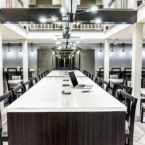 A modern workspace with a long table, chairs, laptops, and lighting fixtures. The space is minimalistic with gray tones and additional seating areas.