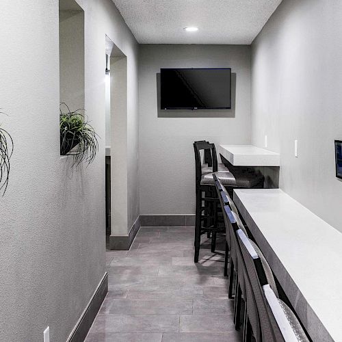 A modern, narrow office space with a long white desk, barstools, a wall-mounted TV, plants, and a small hallway.