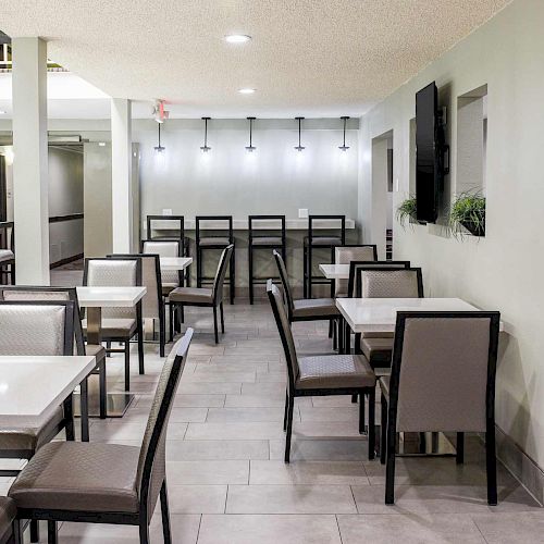 The image shows a modern dining area with tables and chairs, a bar-like counter with high stools, gray flooring, and minimalistic wall decor.