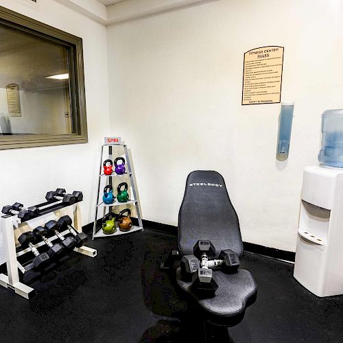 This image shows a small gym corner with a bench, dumbbells, kettlebells, a water cooler, and a hand sanitizer dispenser.