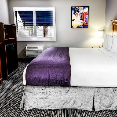 A hotel room with a bed, TV, dresser, and wall art above the bed. The bed has a white comforter and a purple blanket. 