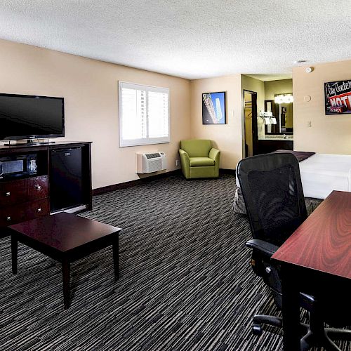 The image shows a modern hotel room with a bed, TV, desk, chair, coffee maker, and armchairs, decorated with artwork on the walls.