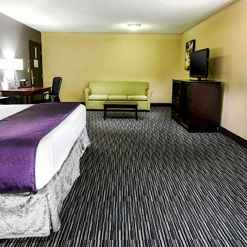 A hotel room with a bed, sofa, TV, desk, and chair. The bed has white and purple bedding, and there is a greenish sofa and chair.