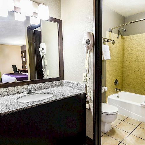 A hotel bathroom with a granite countertop sink, wall-mounted hairdryer, toilet, and bathtub; view of a bedroom with a green sofa in the background.