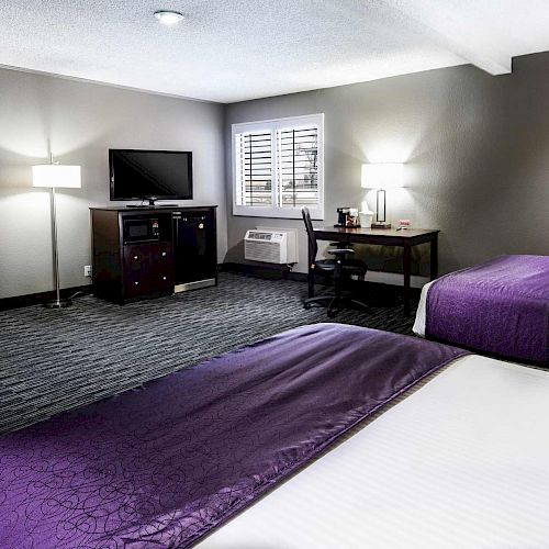 A modern hotel room features two beds with purple runners, a TV, desk with lamp, wall art, green couch, and white walls with gray accents.