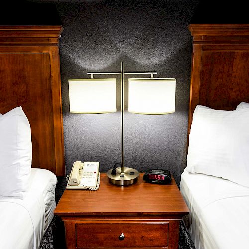 A bedside table with a lamp, phone, and alarm clock sits between two beds with white linens and dark wooden headboards.