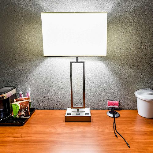 A desk with a lamp, a coffee maker, condiments tray, a phone stand, and a trash bin, creating a tidy workspace setup.