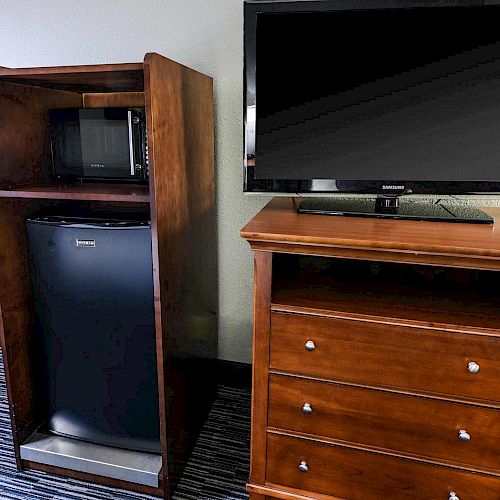 The image shows a wooden shelving unit holding a microwave and a mini refrigerator, next to a wooden dresser with a TV on top.