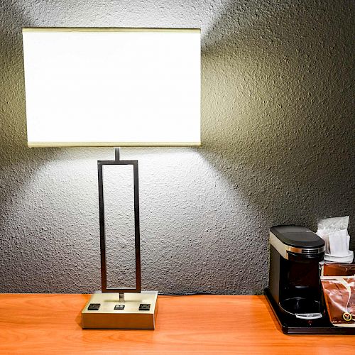 A table with a modern lamp, a coffee maker, coffee pods, stir sticks, and napkins arranged neatly to the right, resting on a wooden surface.