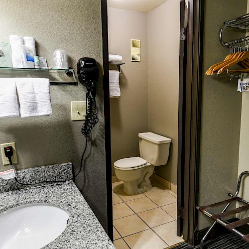 A bathroom with a sink, toiletries on shelves, towels, a wall-mounted hairdryer, a toilet, and a hanging rod with hangers in an adjoining area.