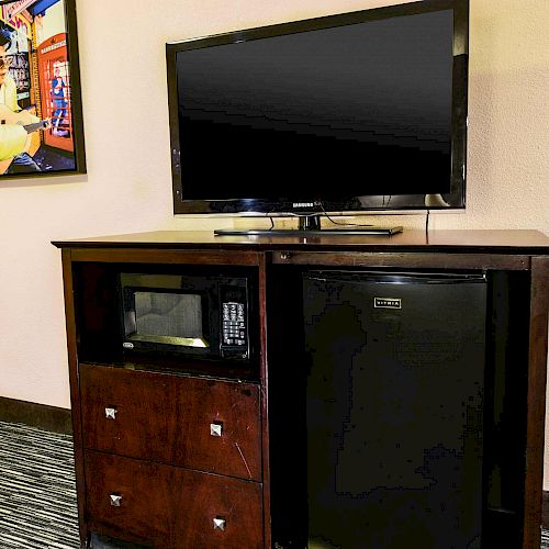 The image shows a television on a dark wood cabinet with a microwave, a mini-fridge, drawers, and wall art. The setup appears in a room.