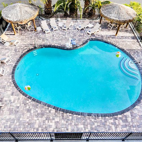 An aerial view of a heart-shaped swimming pool surrounded by lounge chairs, umbrellas, and palm trees, enclosed by a black fence.