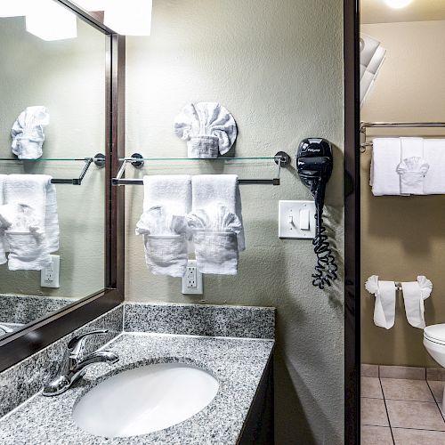 A bathroom with a sink, mirror, towels, a hairdryer, and a toilet, featuring a clean and neatly organized arrangement, with folded towels.